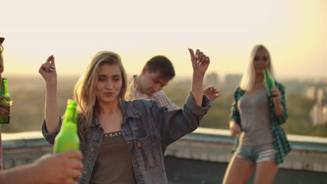 The-young-girl-is-dancing-on-the-roof-with-her-friends-on-the-party.-She-closes-her-eyes-and-enjoys-the-time-in-shorts-and-a-light-denim-jacket-in-summer-everning.-Her-blond-hair-is-flying-in-the-wind.-She-is-dancing-with-her-arms-and-body.